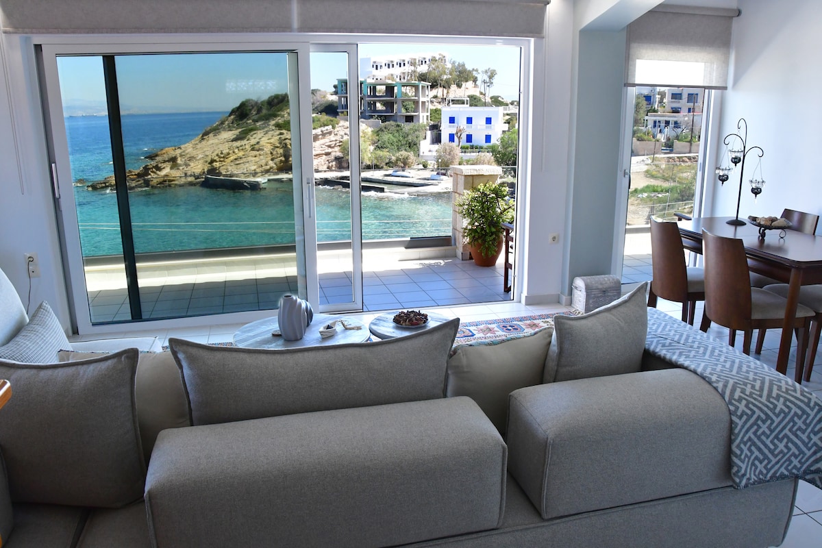 A spacious living area is visible, featuring a large, comfortable sofa and a coffee table. Expansive glass doors frame a stunning sea view, allowing natural light to fill the space. A dining table is positioned nearby, complementing the serene coastal ambiance.