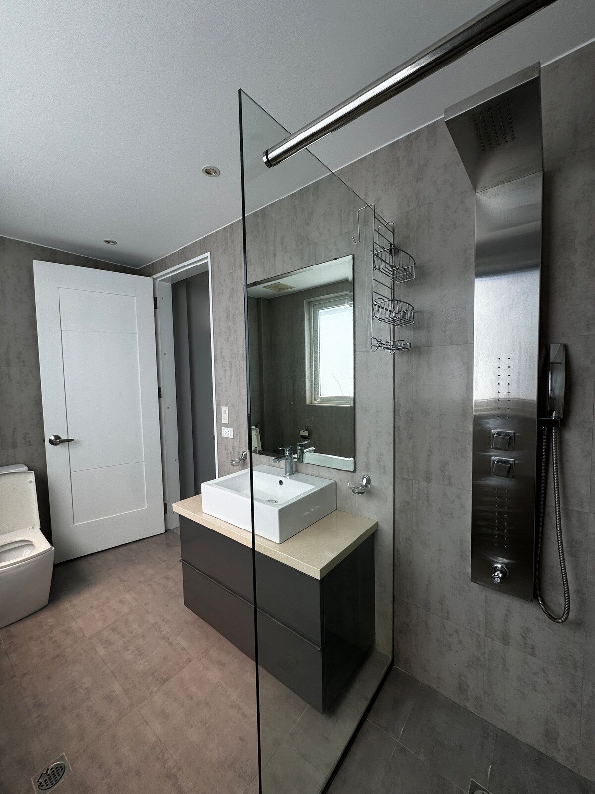 A modern bathroom is presented with a walk-in shower enclosed by clear glass. A sleek vanity with a rectangular sink and minimalist counter space is showcased. Light gray walls and contrasting dark cabinetry enhance the contemporary design. A large mirror reflects the well-lit space.