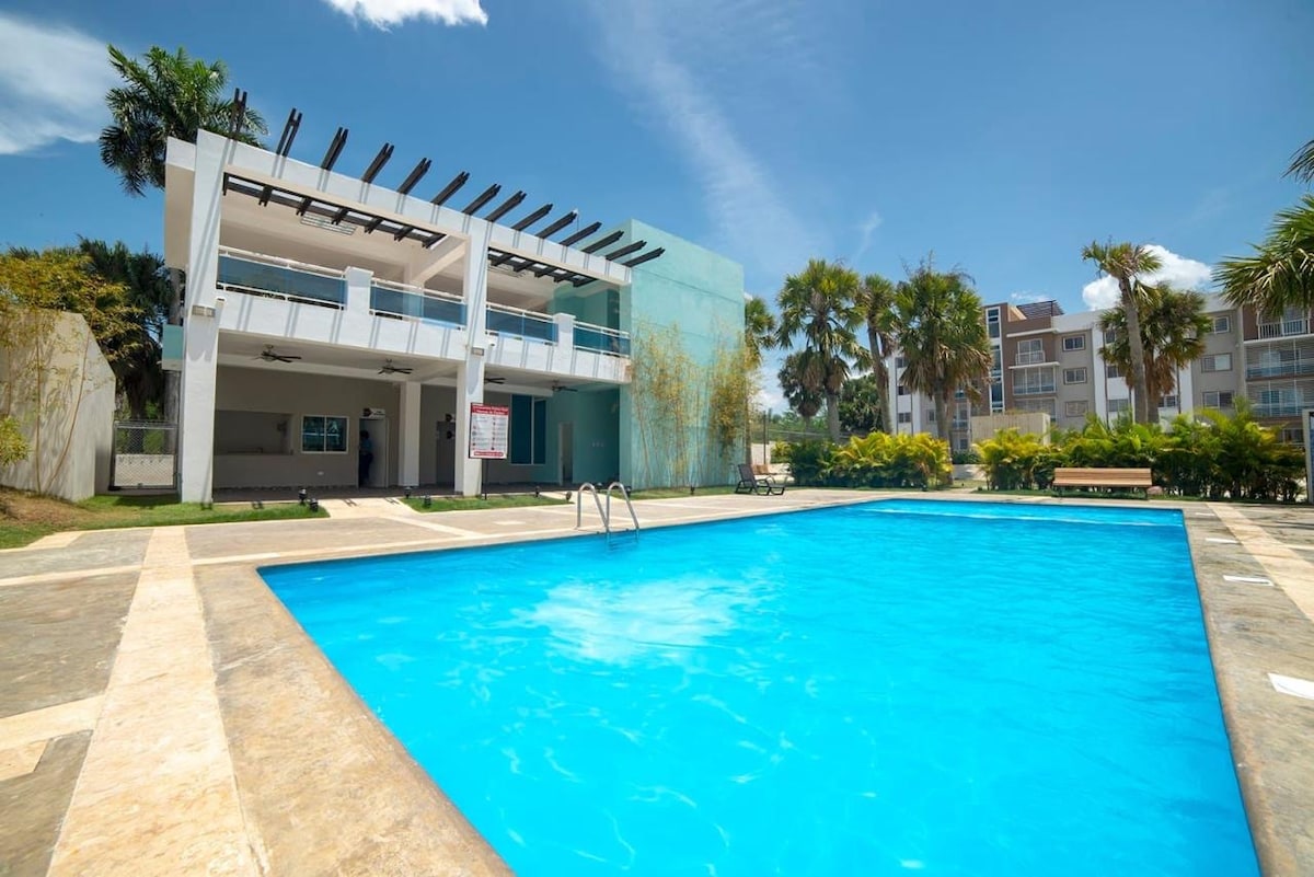 A spacious outdoor area features a large swimming pool surrounded by concrete and grassy landscaping. The building behind the pool has a modern design with large windows and a balcony. Palm trees provide a tropical ambiance, and clear blue skies are overhead.