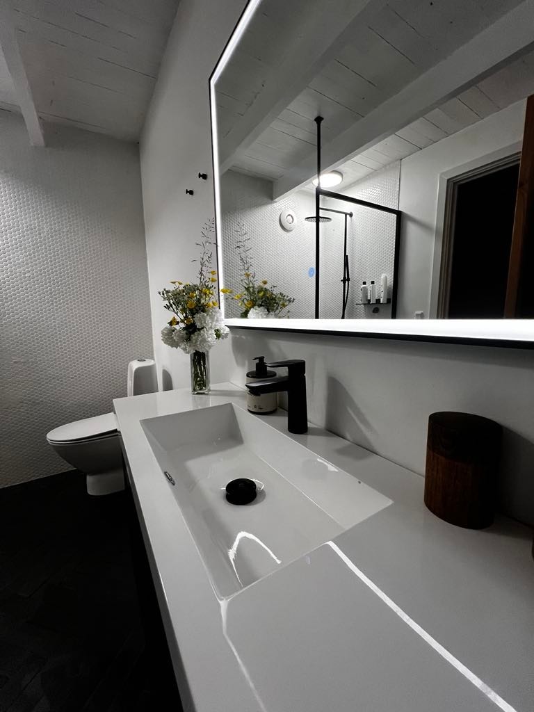 A modern bathroom features a sleek white sink with a single round faucet. The wall-mounted mirror reflects soft lighting. A floral arrangement adds a decorative touch to the countertop, alongside a black soap dispenser and a wooden container. A toilet is visible in the background.