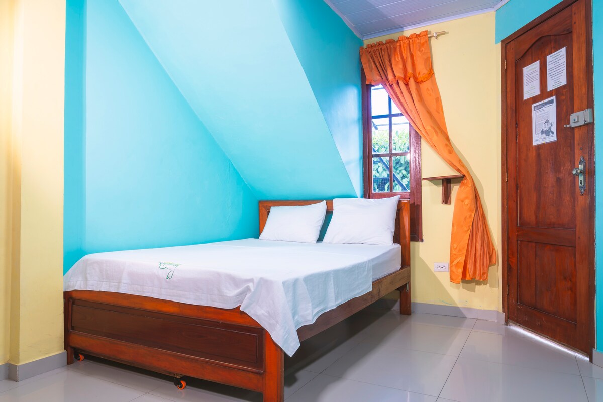 A room features a spacious bed with white linens positioned against a turquoise accent wall. Natural light enters through a window adorned with orange curtains. A wooden door is visible, adding a rustic touch to the inviting space.
