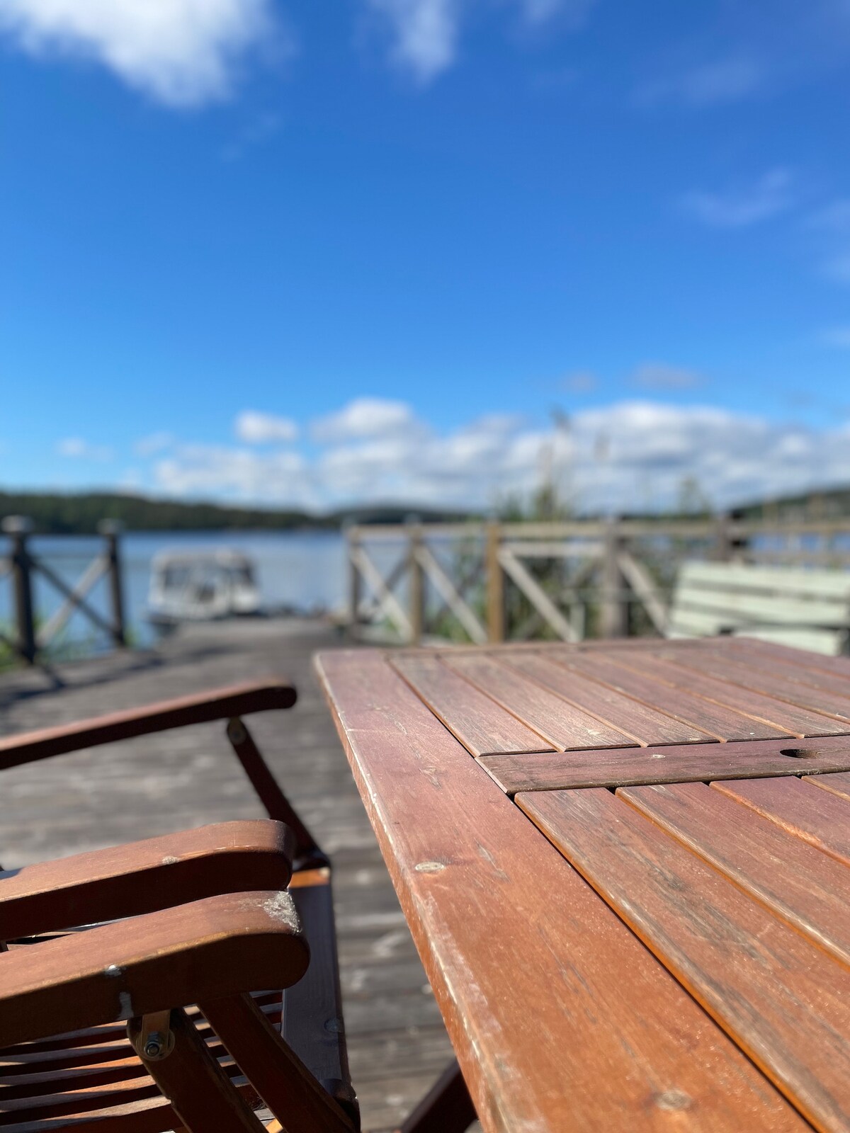A wooden table with a slatted surface is shown alongside a folding chair, both positioned on a deck overlooking a serene body of water. The sky is clear with fluffy white clouds, and a wooden railing outlines the deck.