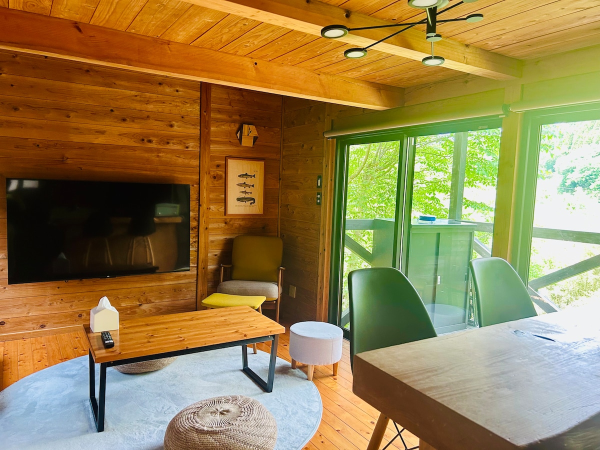 A warm living area presents natural wood paneling with large glass doors providing views of the outdoors. A sizable television is mounted on the wall, while a cozy armchair and a wooden coffee table complement the space. Bright light streams in, enhancing the inviting ambiance.