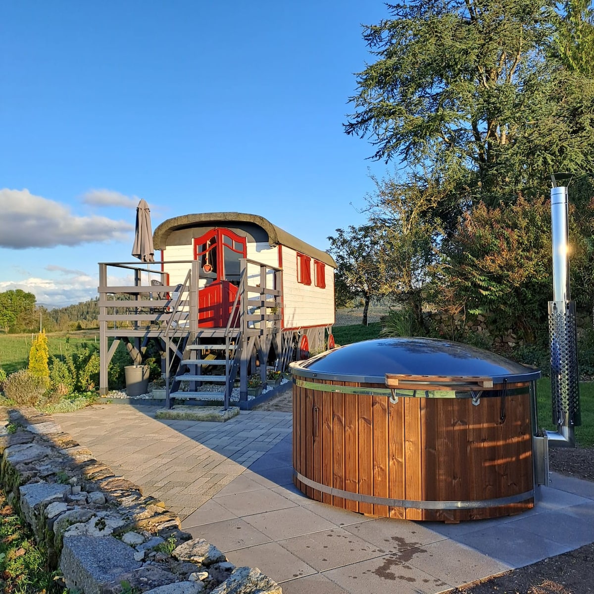 A classic gypsy caravan is positioned on a wooden deck, featuring red accents and a charming staircase. Nearby, a round wooden hot tub is placed on a stone patio, surrounded by greenery and a serene natural landscape.