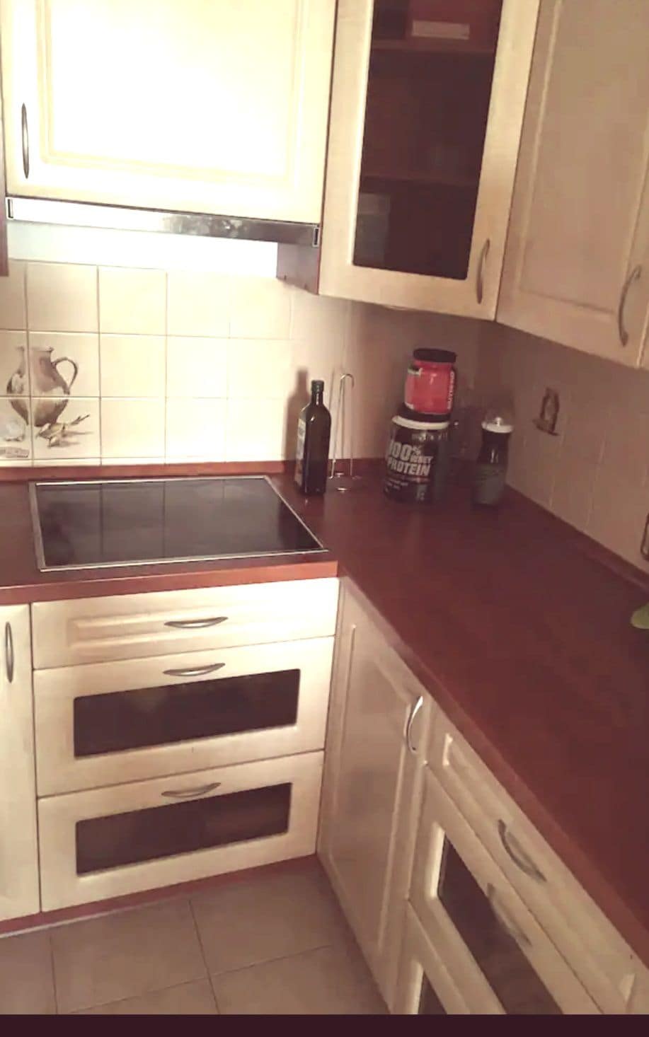 A compact kitchen is presented with light-colored cabinetry and a smooth countertop. Cooking appliances, including a stovetop, are visible. Storage is provided through both upper and lower cabinets. Culinary accessories such as a bottle of oil and a blender are positioned on the counter.