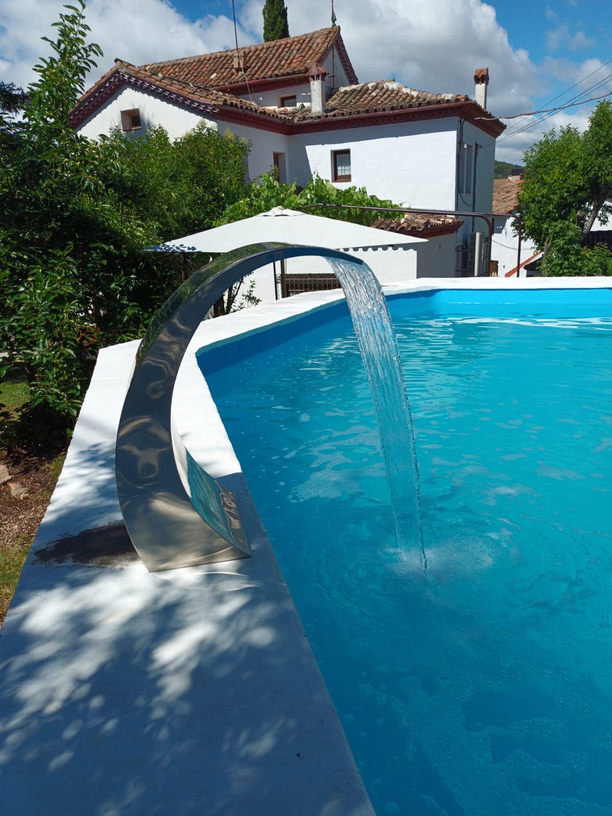 A private swimming pool is displayed with a modern water feature creating a gentle waterfall effect. The pool is surrounded by lush greenery and a charming white building with a terracotta roof is visible in the background, adding to the serene atmosphere.