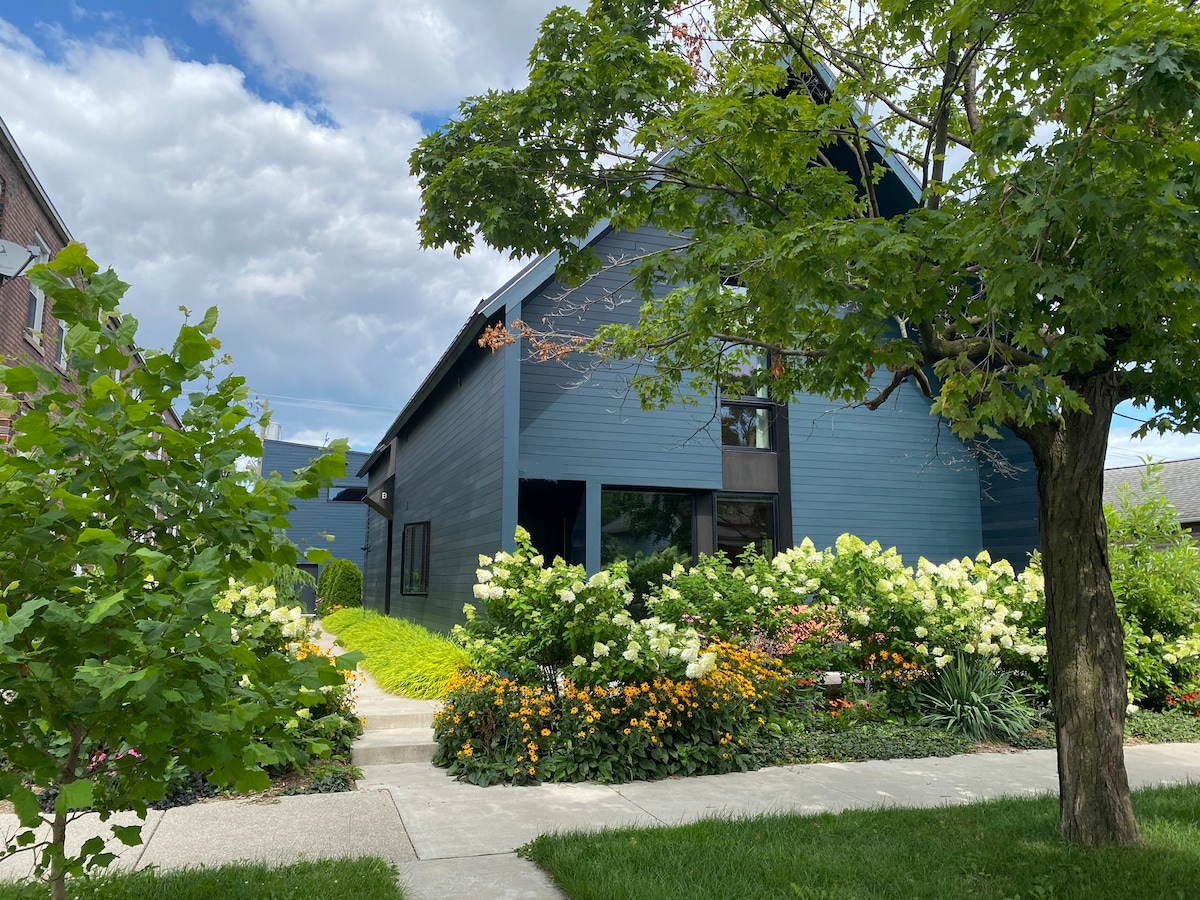 The modern home is surrounded by vibrant greenery and flowering shrubs, including hydrangeas and other colorful blooms. A lush lawn leads to a pathway, inviting guests toward the entrance. The facade features large windows, offering a glimpse into the interior space.