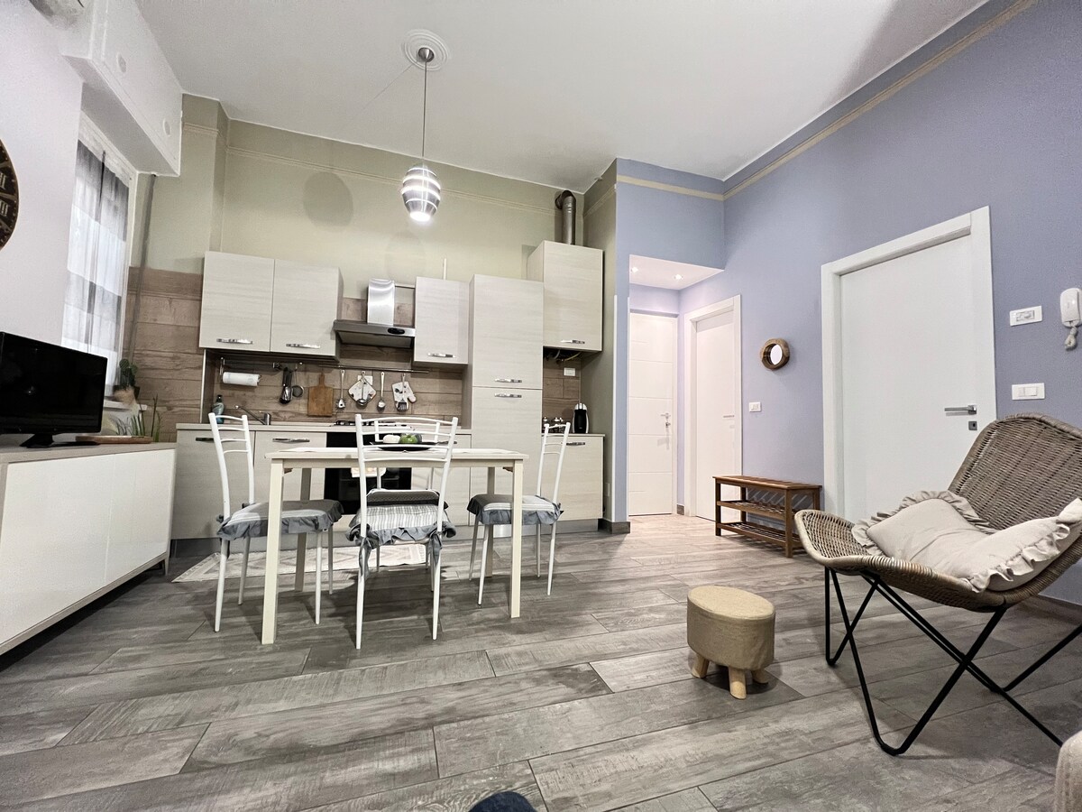 A modern kitchen and dining area is depicted, featuring a light-colored table surrounded by chairs. A sleek kitchen with neutral cabinetry and appliances is visible in the background. The floor is finished in wood-like tiles, and natural light illuminates the space through a window.