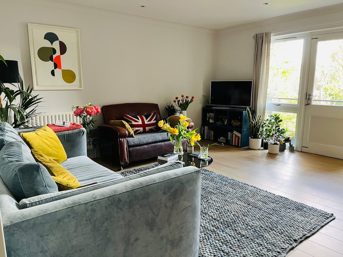 A spacious living area is filled with natural light, featuring a gray sofa and a brown leather armchair. A coffee table with fresh flowers sits at the center. Decorative plants are arranged throughout, and a television is positioned across from the seating. Artwork adorns the wall.