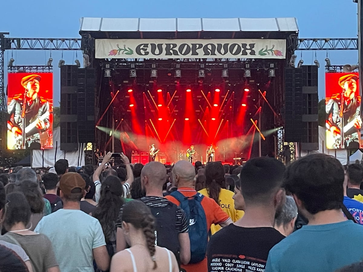 An outdoor concert scene is captured, showcasing a crowd of diverse individuals enjoying a live performance. Bright red stage lights illuminate three musicians on a large stage, with large screens displaying their images. The atmosphere is vibrant, filled with the energy of the audience.