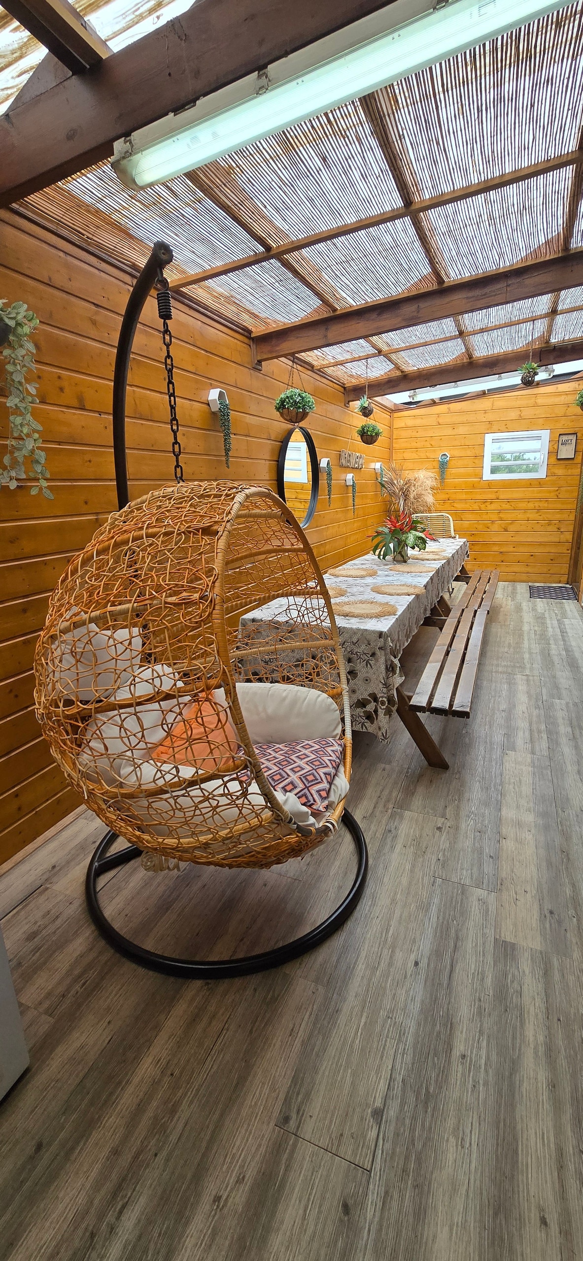 A cozy interior space features a swinging chair made of woven material, accompanied by a long dining table set for meals. The room is enhanced by wooden walls and a series of potted plants, providing a warm and inviting environment.