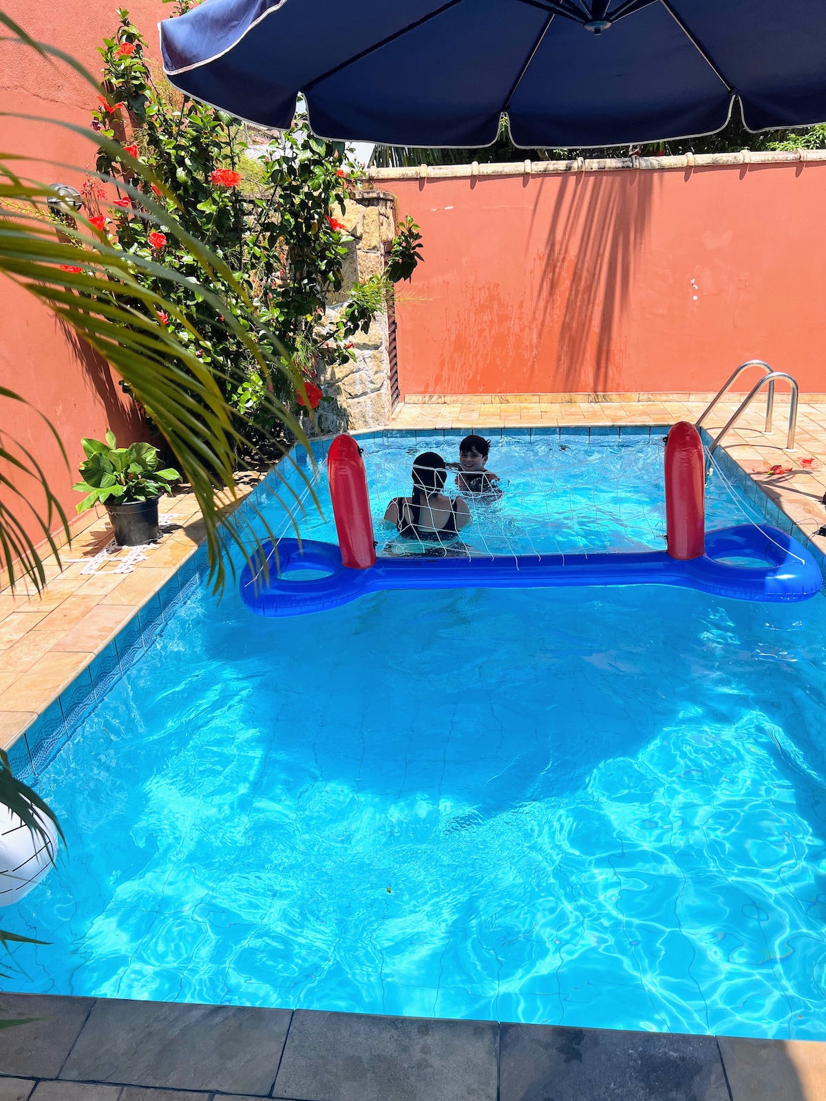 A clear and inviting pool is featured, showcasing vibrant blue water glistening under sunlight. Two individuals are seen enjoying the water, surrounded by lush greenery and a red wall. A large umbrella provides shade, and a pool safety barrier is visible.