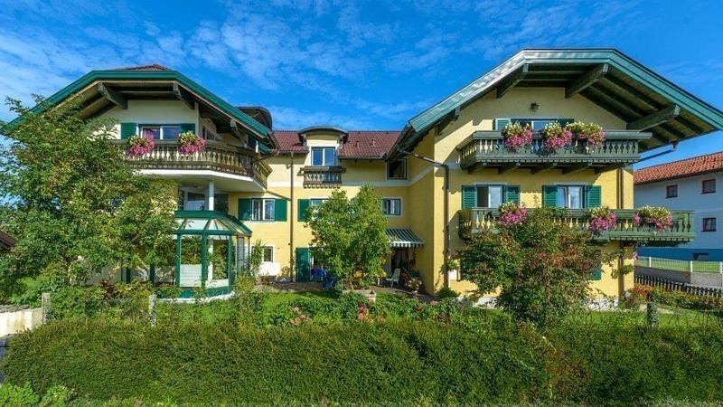 A charming apartment building is set against a clear blue sky, featuring a vibrant yellow exterior with balconies adorned with colorful flowers. Lush greenery surrounds the property, creating a welcoming environment. A gazebo is visible in the garden area, offering a spot for relaxation.