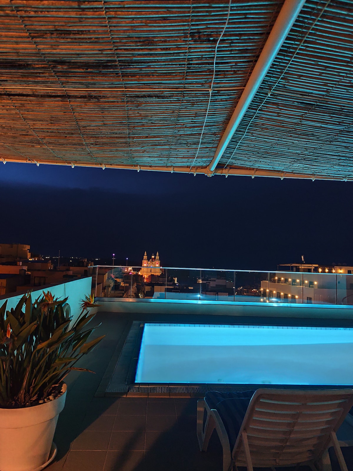 The rooftop pool area is illuminated gently, showcasing a clean swimming pool. A lounge chair rests nearby, and decorative greenery emerges from a planter. The distant cityscape is visible under a night sky, with twinkling lights adding to the tranquil setting.