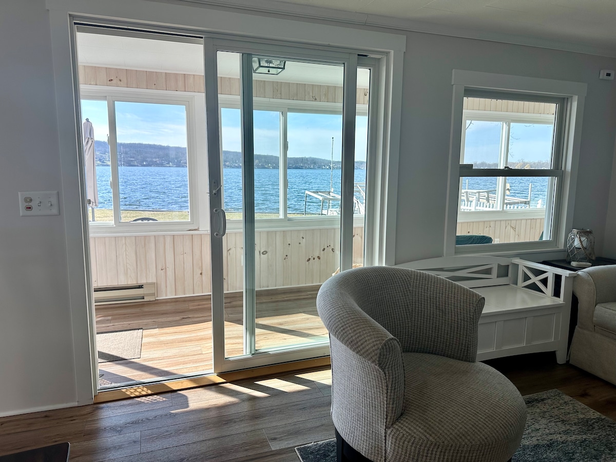 A glass sliding door opens to a screened porch, providing an unobstructed view of the lake. The light wood paneling of the porch adds warmth, while the sunlight illuminates the inviting indoor seating area, creating a bright and airy ambiance.