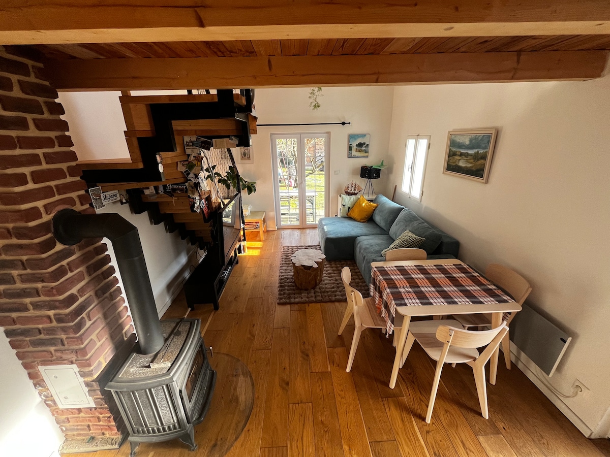 An open loft space features wooden flooring and a cozy living area with a soft sofa. A small dining table with light-colored chairs is positioned nearby. A wood-burning stove adds warmth, while glass doors lead to the outdoor area, allowing natural light to fill the room.