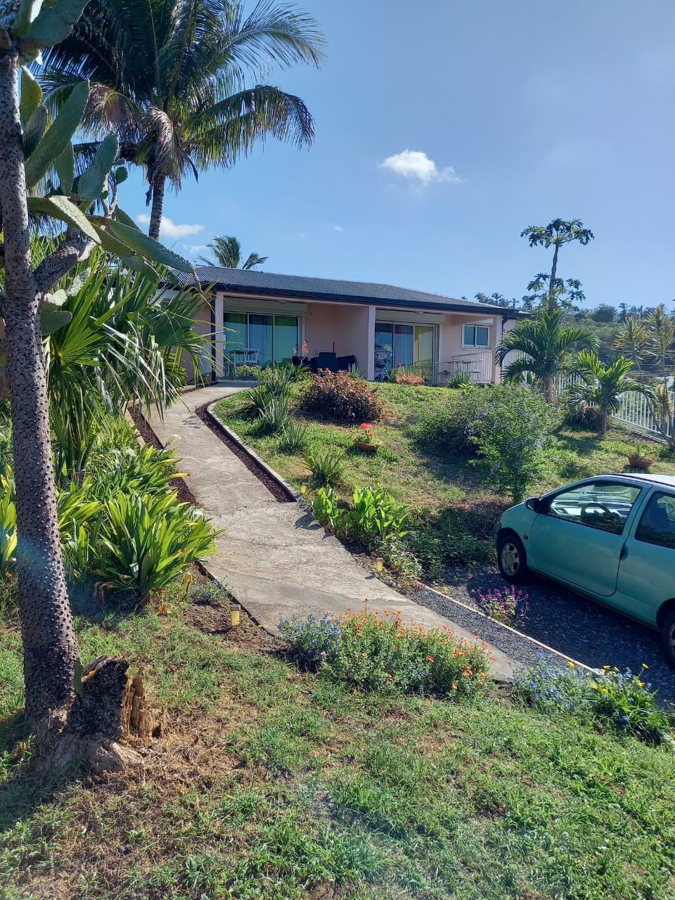 A residence is set amidst lush tropical landscaping, featuring a winding pathway leading to a light-colored building. Palm trees and vibrant flowers enhance the exterior, while a parked car is visible nearby. The structure is complemented by a clear blue sky overhead.