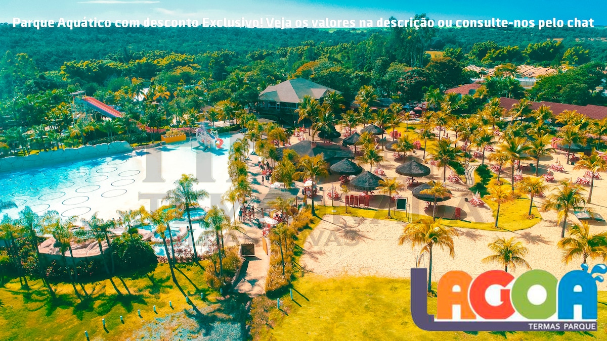 A vibrant water park features a large swimming area, surrounded by various thatched umbrellas and lounge chairs. The layout includes an expansive pool with clear blue water, and lush greenery lines the background, creating a pleasant recreational space.