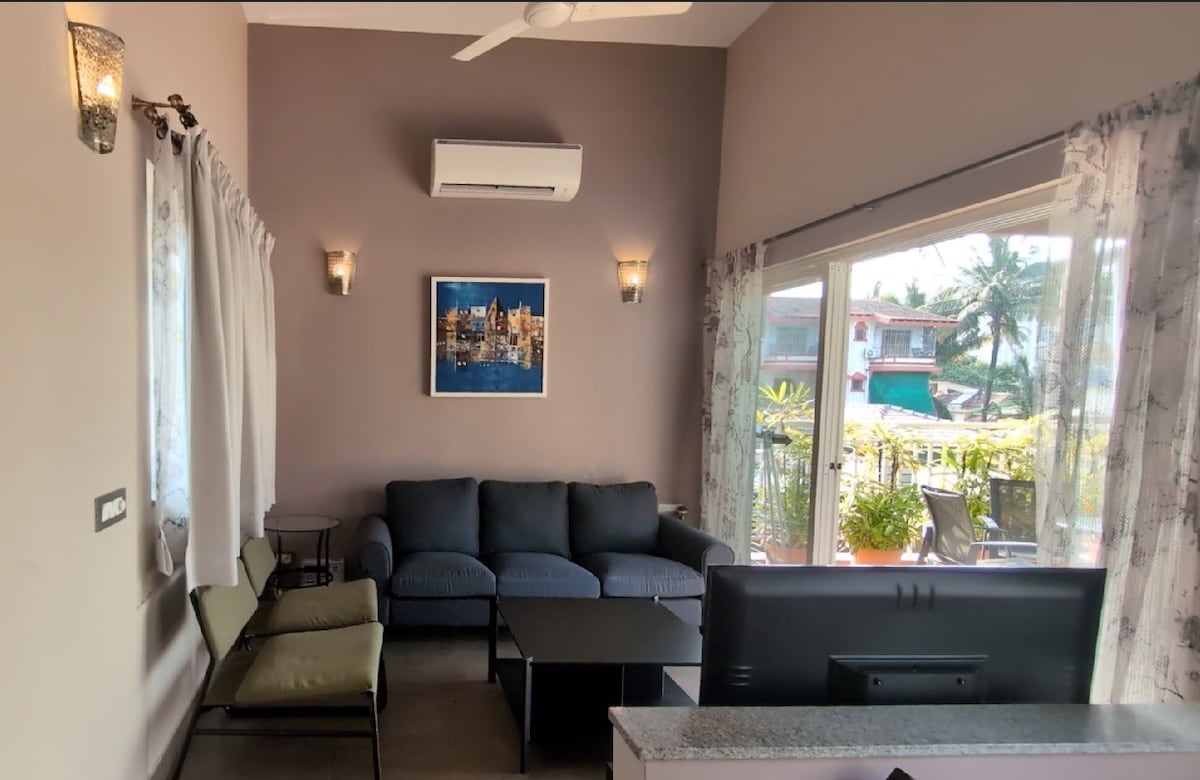 The cozy living area is furnished with comfortable sofas and a low coffee table. Natural light filters through large glass doors, leading to a terrace, while an air conditioning unit ensures a pleasant atmosphere. A wall-mounted television provides options for entertainment.