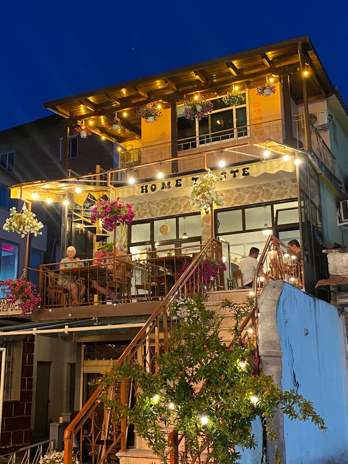A two-story building is illuminated by warm lighting at night, featuring a welcoming outdoor terrace adorned with colorful flowers. The name 'HOME TASTE' is prominently displayed, and diners can be seen enjoying their meals through the large windows, creating a lively atmosphere.