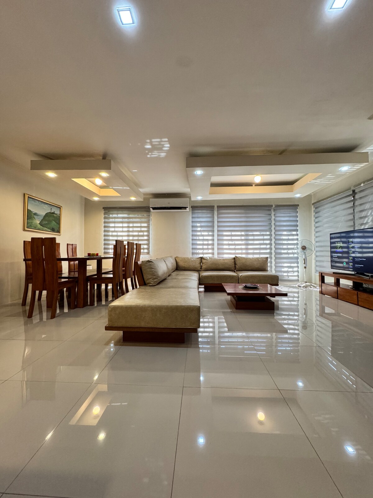 The living and dining area features polished tile flooring and high-end hardwood furniture. A spacious sofa faces a low coffee table, with a dining set positioned nearby. Large windows are fitted with sheer and solid blinds, allowing natural light into the room.