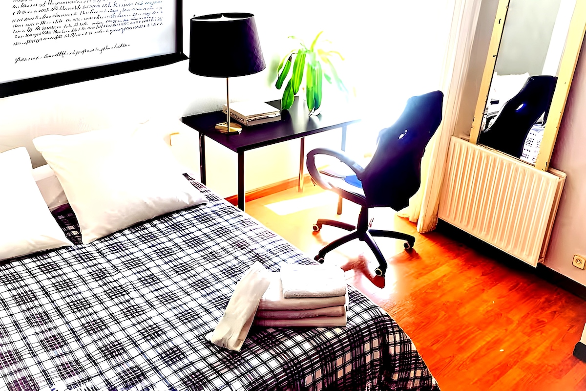 מיטה זוגית הפונה למראה גדולה ושולחן עם תאורה טבעית. רצפת עץ, ריהוט מינימליסטי והרמוניה בסידור החלל.