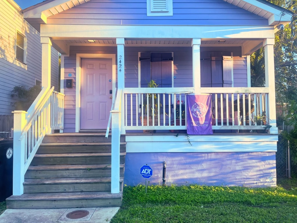 A charming camel-back home features a welcoming front porch with white railings and a series of steps leading up. The lavender exterior is complemented by light purple shutters. A flag hangs gracefully, while a security sign is positioned on the grass.