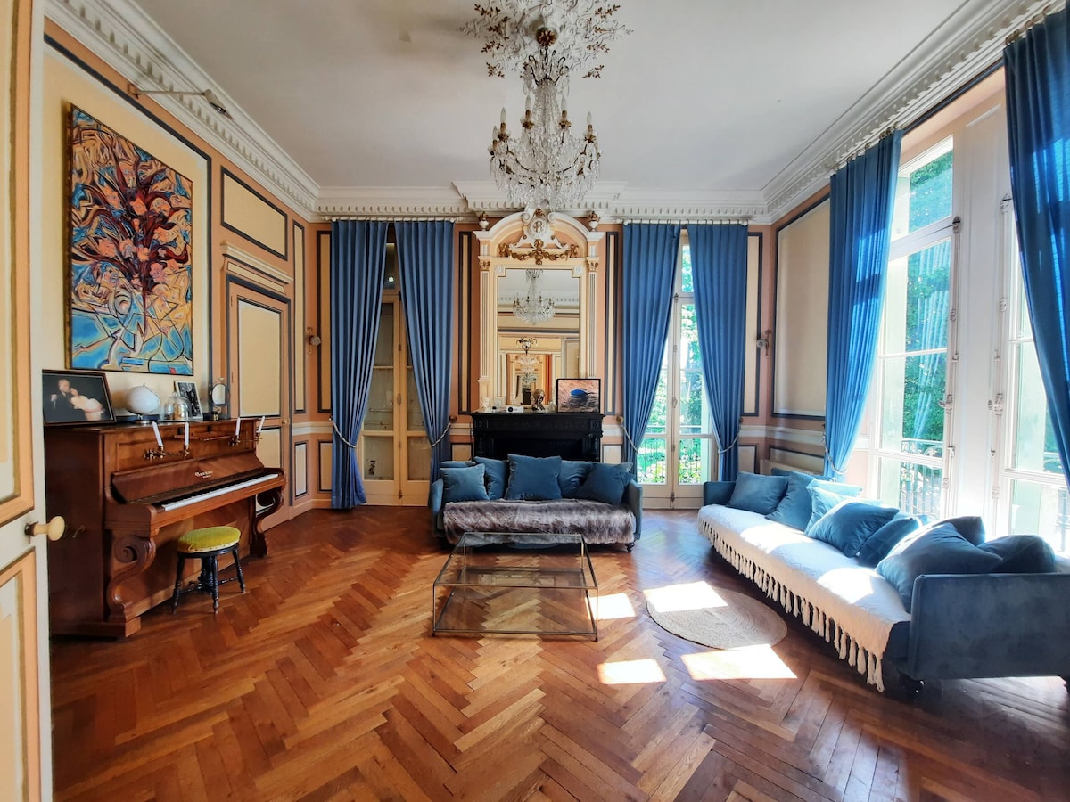 A spacious living room features high ceilings and elegant molding. Large windows dressed with blue curtains allow natural light to filter in, illuminating polished wooden floors. A grand piano is visible beside a plush sectional sofa, and decorative mirrors add depth to the room.