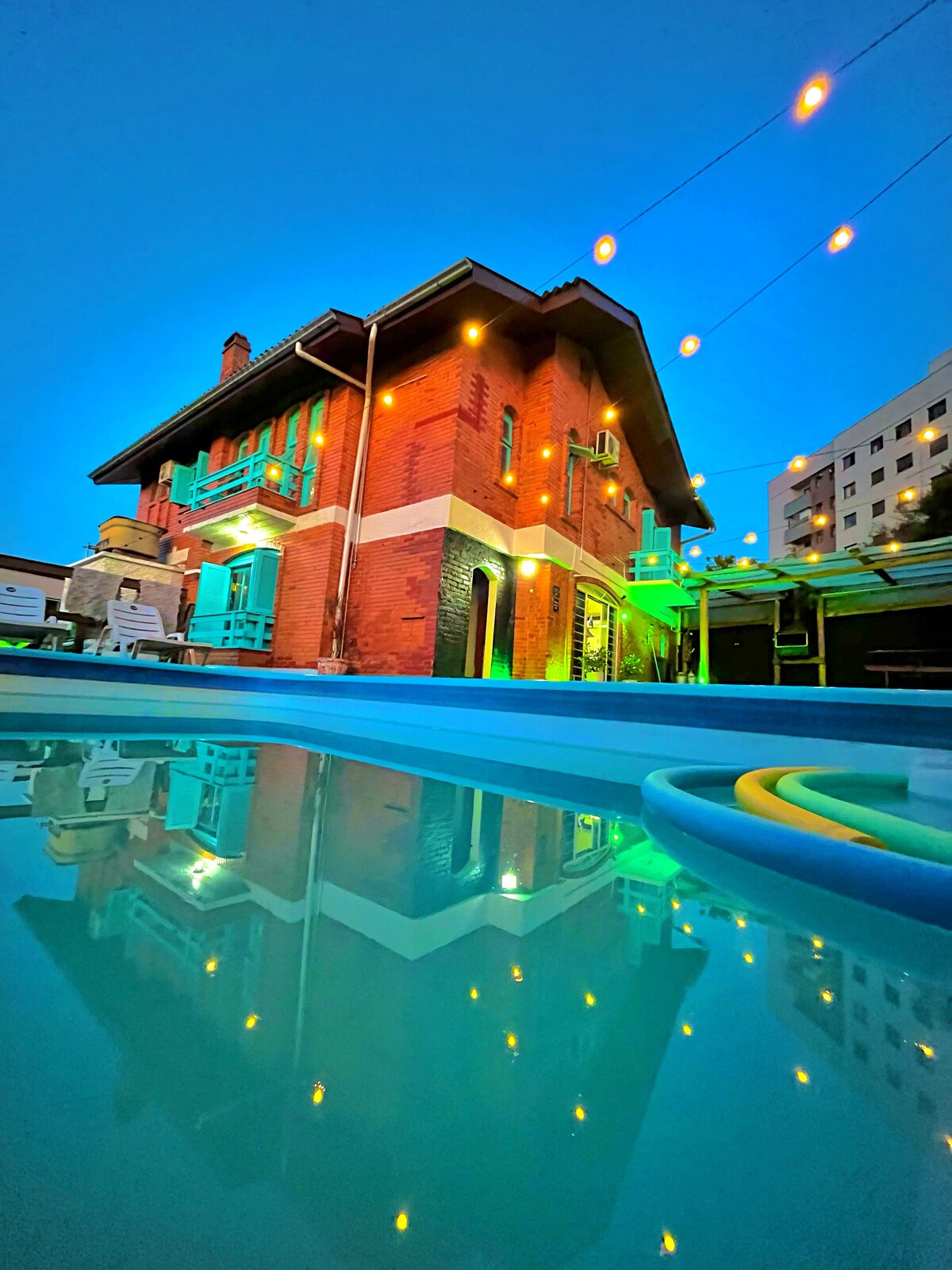 The exterior of a rustic-style house is illuminated in warm tones against a twilight sky. Reflections can be seen in the pool's calm water. Decorative string lights are draped across the outdoor area, adding to the inviting ambiance.
