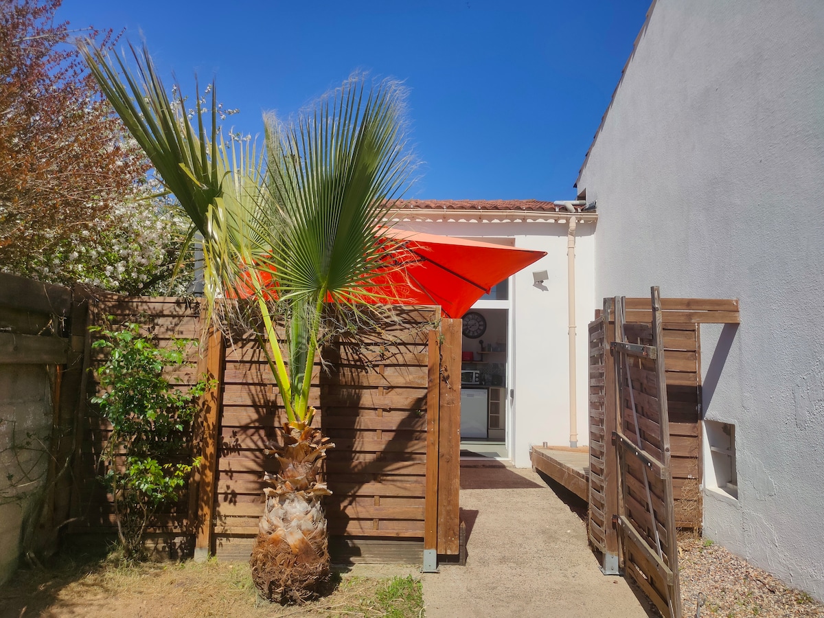 A bright entrance is framed by a lush palm tree and wooden fencing. A vibrant red awning provides shade, leading into a well-lit area that suggests access to the interior. The surrounding greenery adds to the inviting outdoor space.