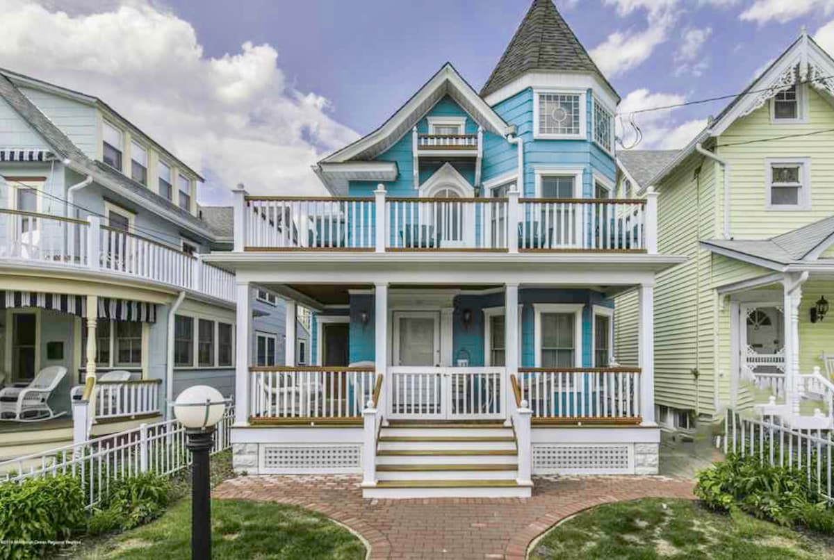 A charming Victorian beach house is showcased, featuring a distinct blue exterior and a prominent pointed turret. A spacious front porch with white railings invites relaxation, and a brick walkway leads to the entrance. Nearby houses exhibit similar coastal architectural styles.