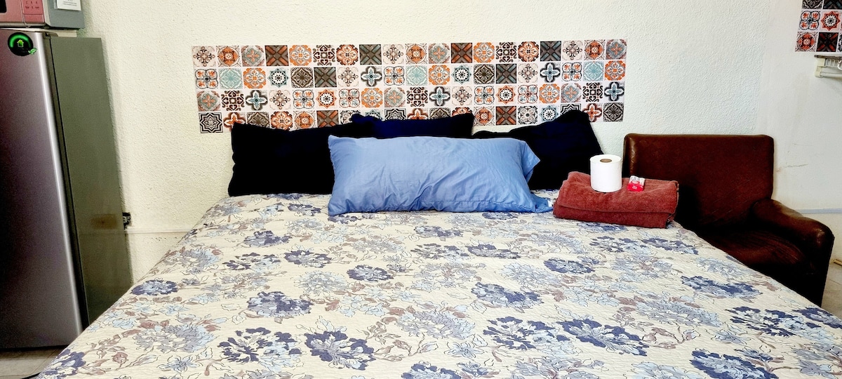 A spacious bed is featured with a floral-patterned cover and complemented by multiple pillows in blue and dark tones. A small brown chair is positioned nearby, with rolled towels placed neatly on the bed. A decorative tiled backsplash adds character to the sleeping area.