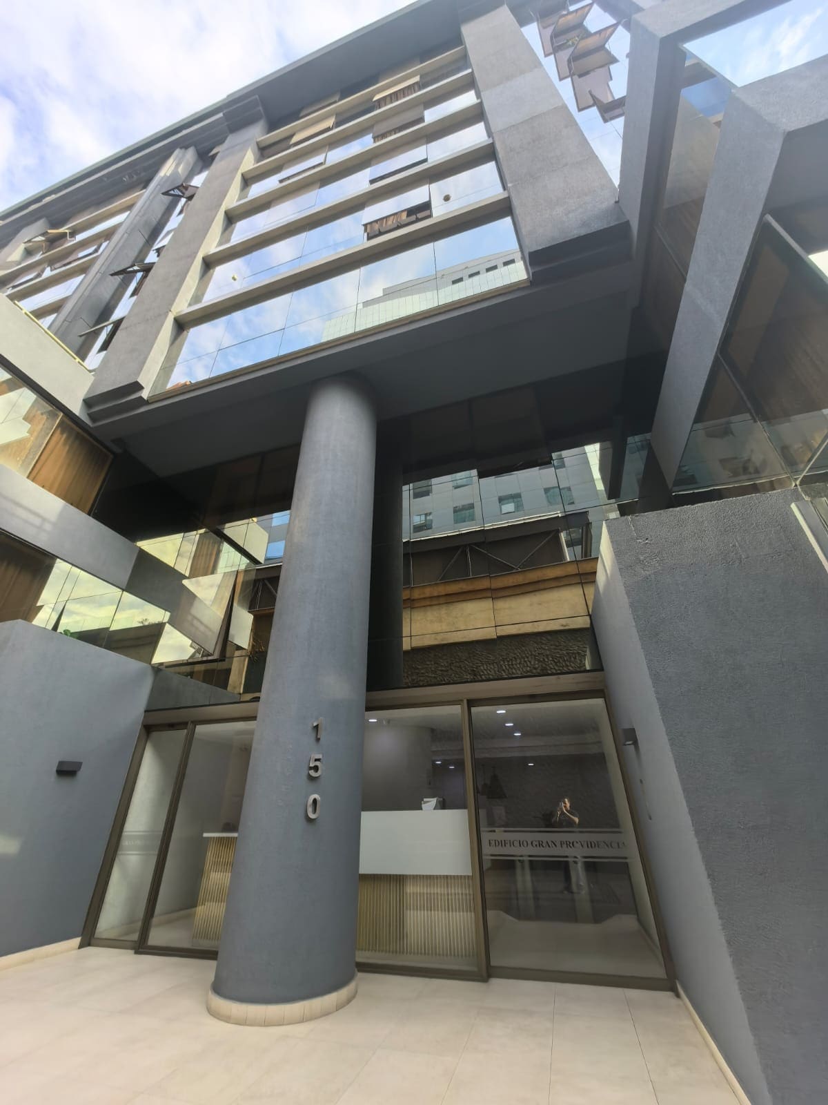 The entrance of the building is marked by a tall, grey column supporting the upper structure. Glass doors provide access to the lobby area, revealing a modern interior with a reception desk. Large windows allow for ample natural light, emphasizing the contemporary design.
