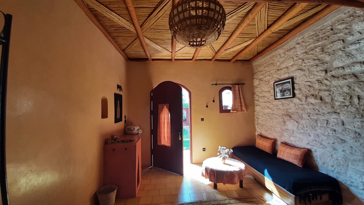 A cozy entryway features a stone wall along one side and a wooden ceiling with exposed beams. A dark wooden door leads outside, while a low wooden table is placed on the tiled floor. A long cushion lines the wall, offering seating.