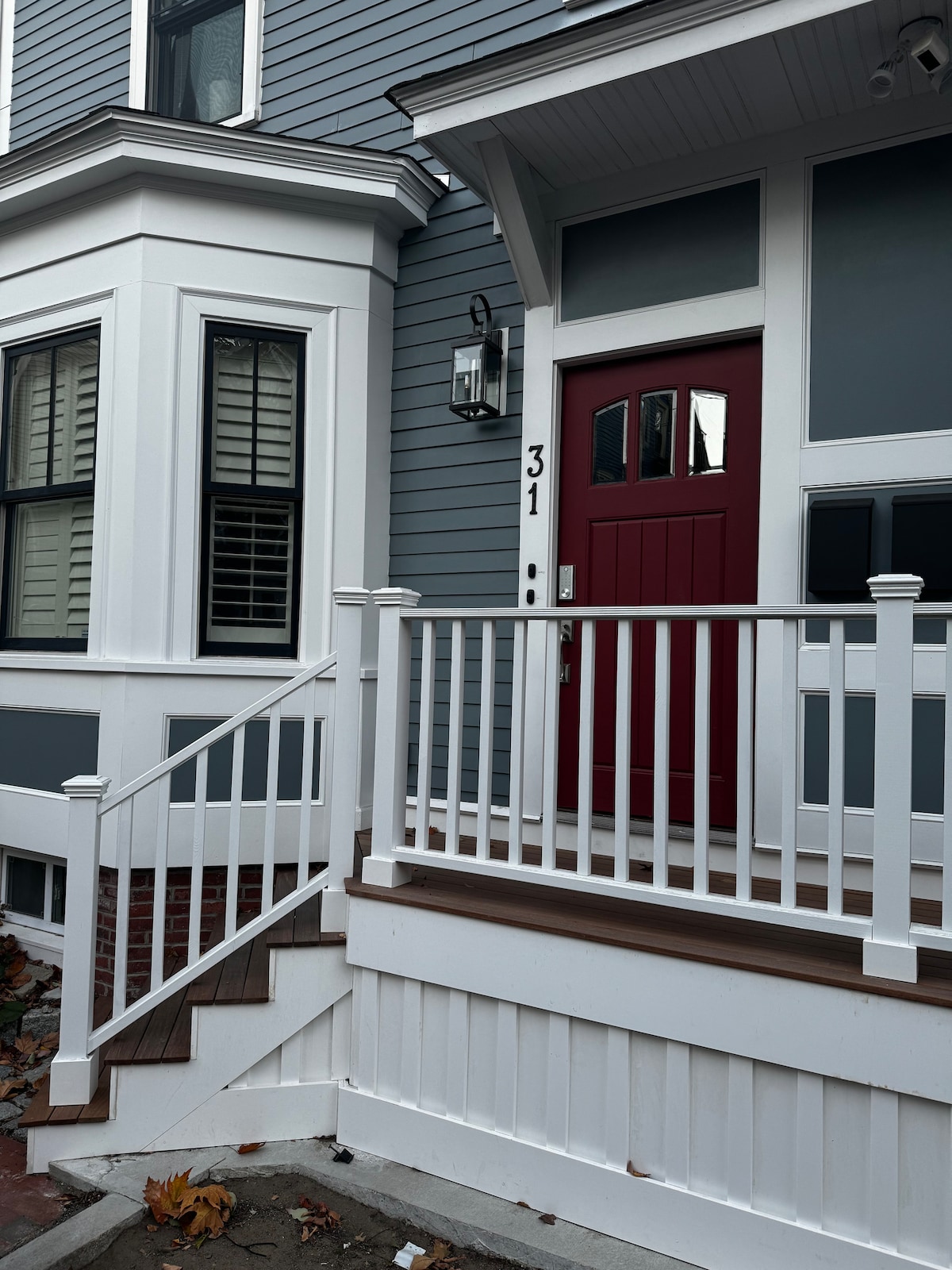 The image shows a well-maintained entrance featuring a red front door complemented by white trim. A set of stairs leads up to the door, flanked by a railing. The house displays a gray exterior with white accents and windows adorned with shutters.