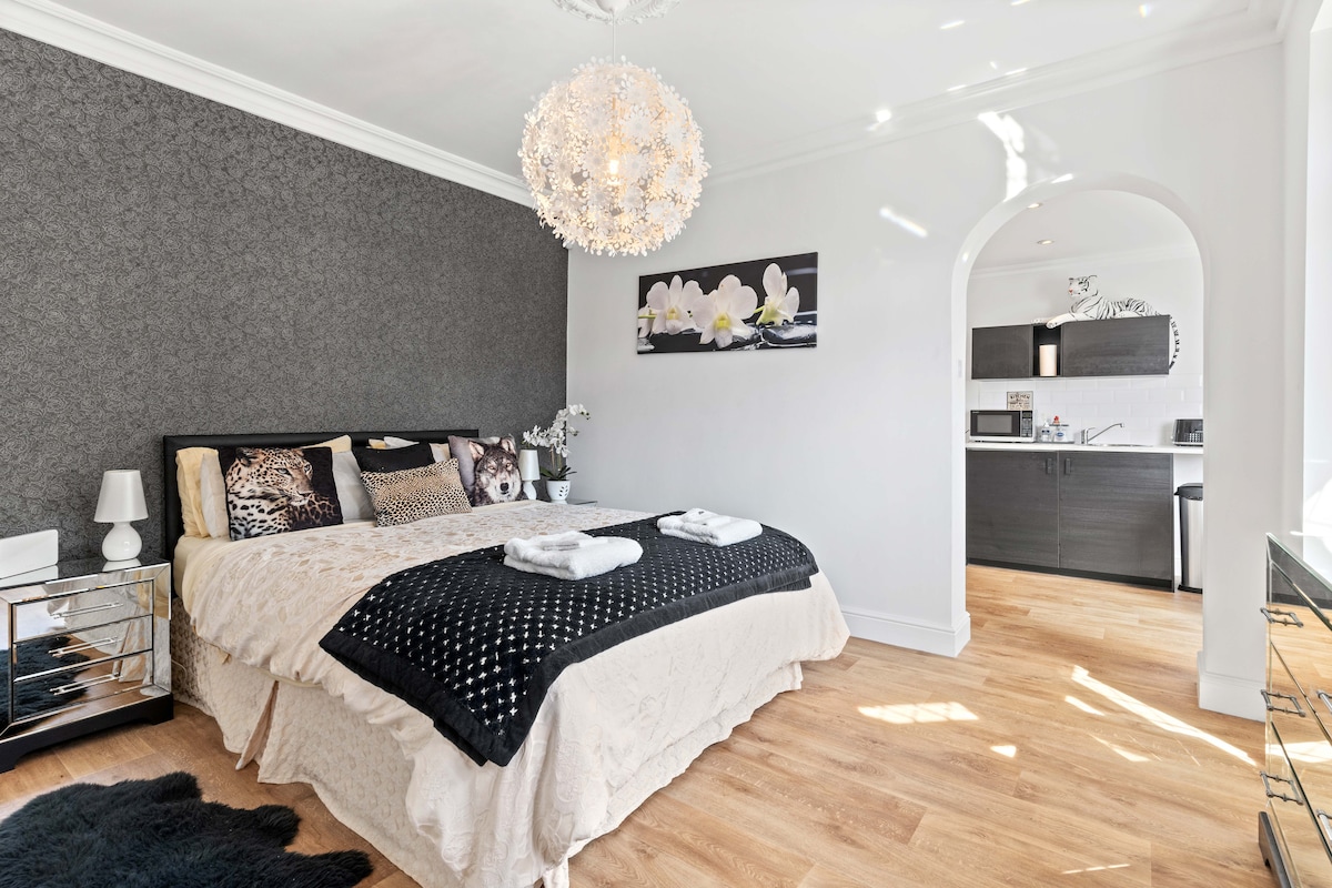 A spacious sleeping area features a king-sized bed elegantly dressed with neutral bedding and decorative pillows. A stylish chandelier provides soft lighting. A kitchenette is visible in the background, equipped with modern appliances. Light floods in through a nearby window, illuminating the warm wood flooring.