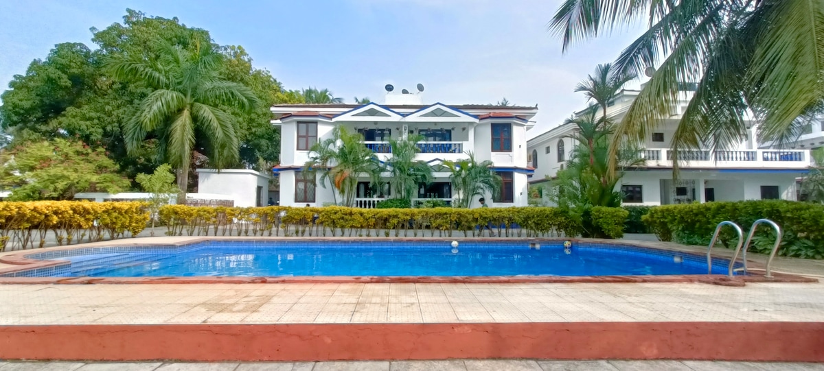 An inviting swimming pool is framed by lush greenery and vibrant hedges, reflecting the surrounding palm trees. In the background, a two-story villa is visible with multiple balconies, blending into the tranquil setting of the property.