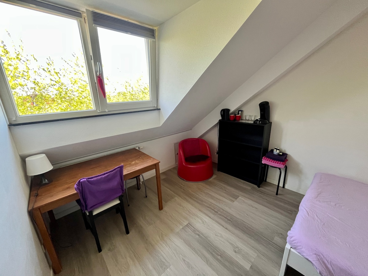 A well-lit room features an angled ceiling and large windows that allow natural light to enter. A wooden desk with a chair, a compact bookshelf, and a red accent chair are present. A small table is arranged for beverages, offering functionality and comfort.