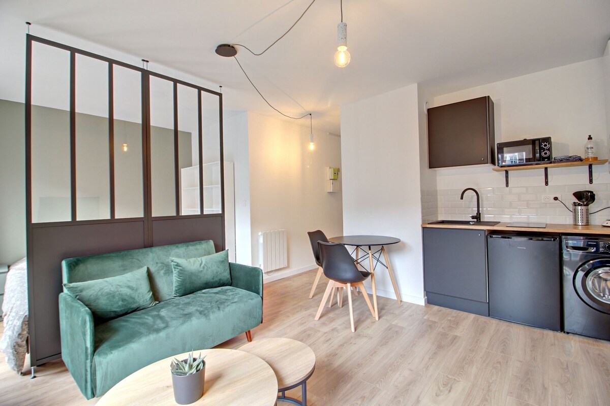 An open living space features a green sofa positioned across from a kitchen area with modern cabinetry. A round dining table for two sits nearby, while the kitchenette is equipped with a microwave, stovetop, and washing machine. Soft lighting enhances the ambiance.
