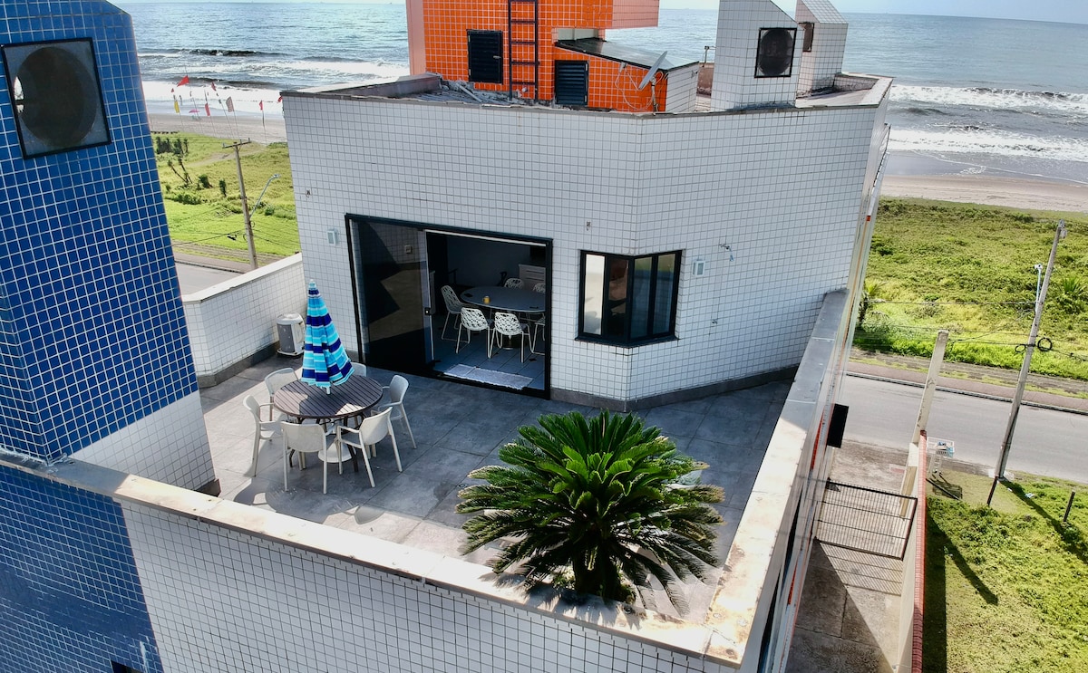An outdoor terrace features a dining area with chairs and a table, surrounded by a clean, spacious surface. A solitary palm plant adds a touch of greenery, while a glimpse of the ocean can be seen in the background, enhancing the coastal ambiance.