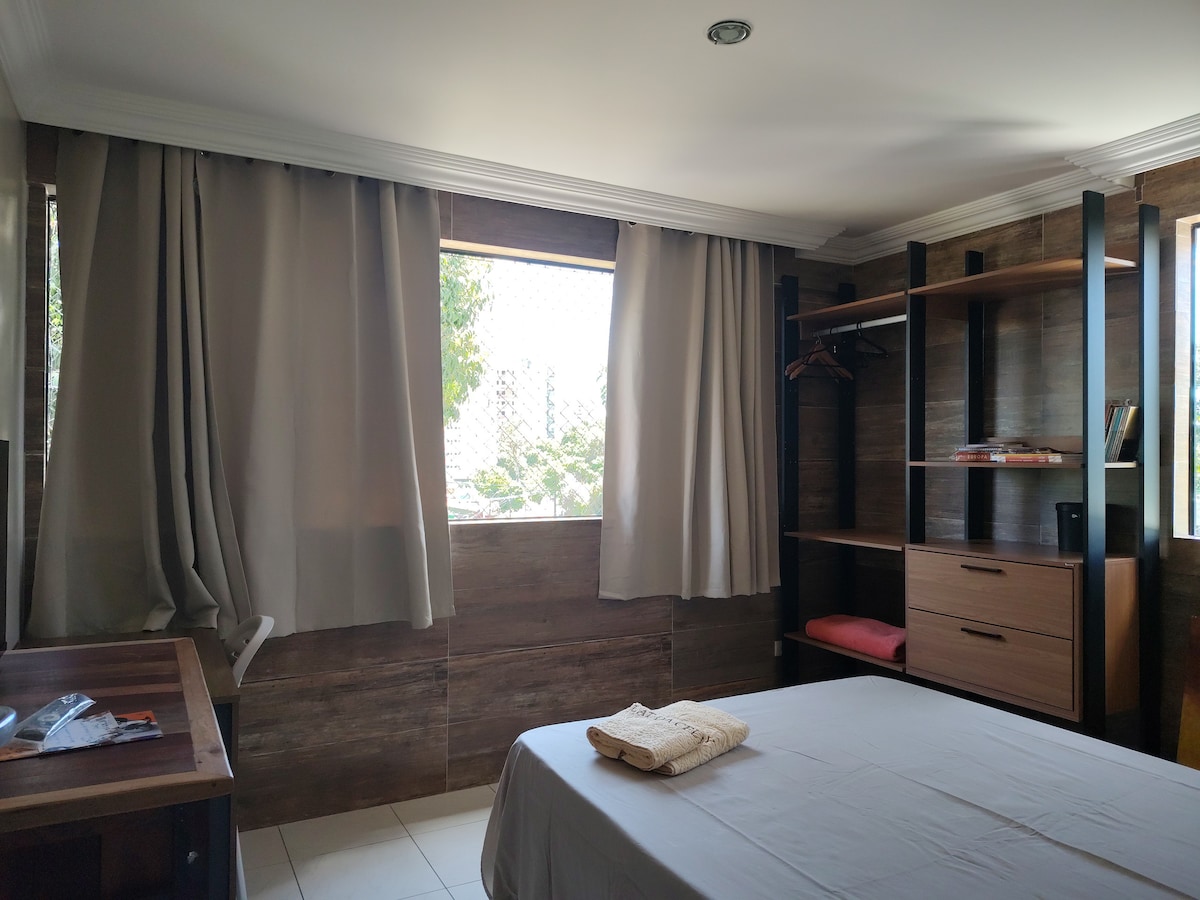 A serene bedroom is showcased, featuring large windows dressed with light-colored curtains. A spacious bed is positioned against a wooden accent wall, and a sleek bookshelf displays books and a few decorative items. A small workspace is visible, enhancing the room's functionality.
