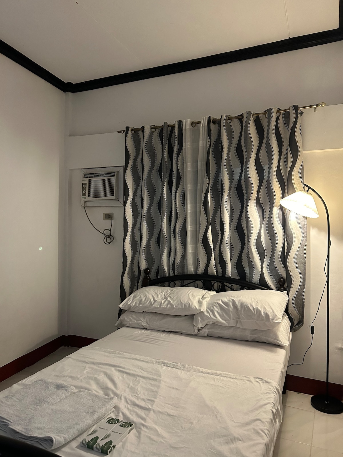 A compact sleeping area is furnished with a single bed, covered with crisp white linens. A decorative curtain with wavy patterns frames a window, while a standing lamp provides additional lighting. An air conditioning unit is mounted on the wall for climate control.
