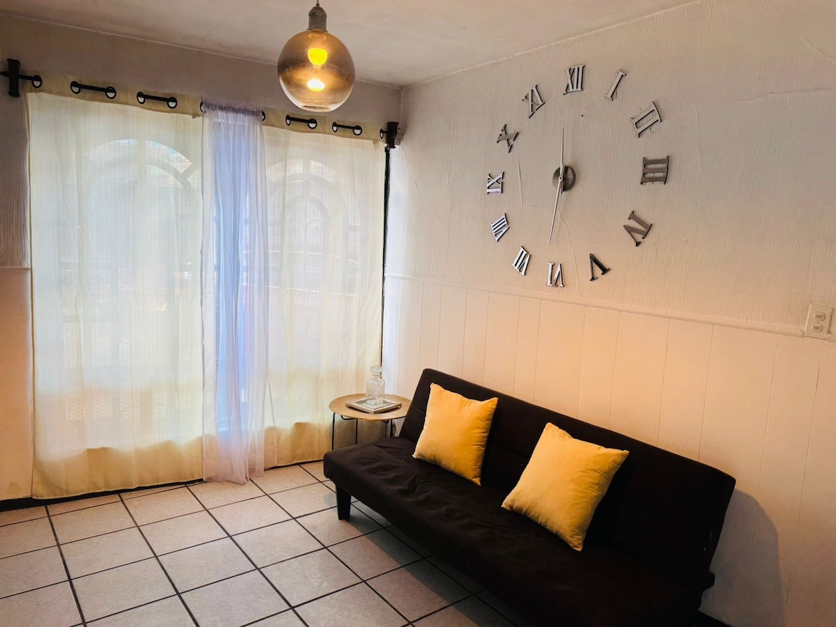 A cozy seating area is presented, featuring a dark sofa adorned with two yellow cushions. A round table is placed beside it, and natural light filters through sheer curtains, illuminating the space. A large wall clock with Roman numerals is mounted above the sofa.