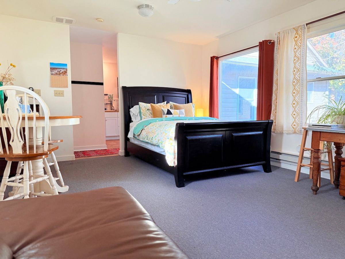 A spacious studio layout is revealed, featuring a large black bed with a colorful quilt and decorative pillows. Natural light filters in through expansive windows with light curtains. A wooden dining table and chairs are visible in the foreground, complemented by a cozy seating area with a leather sectional couch.