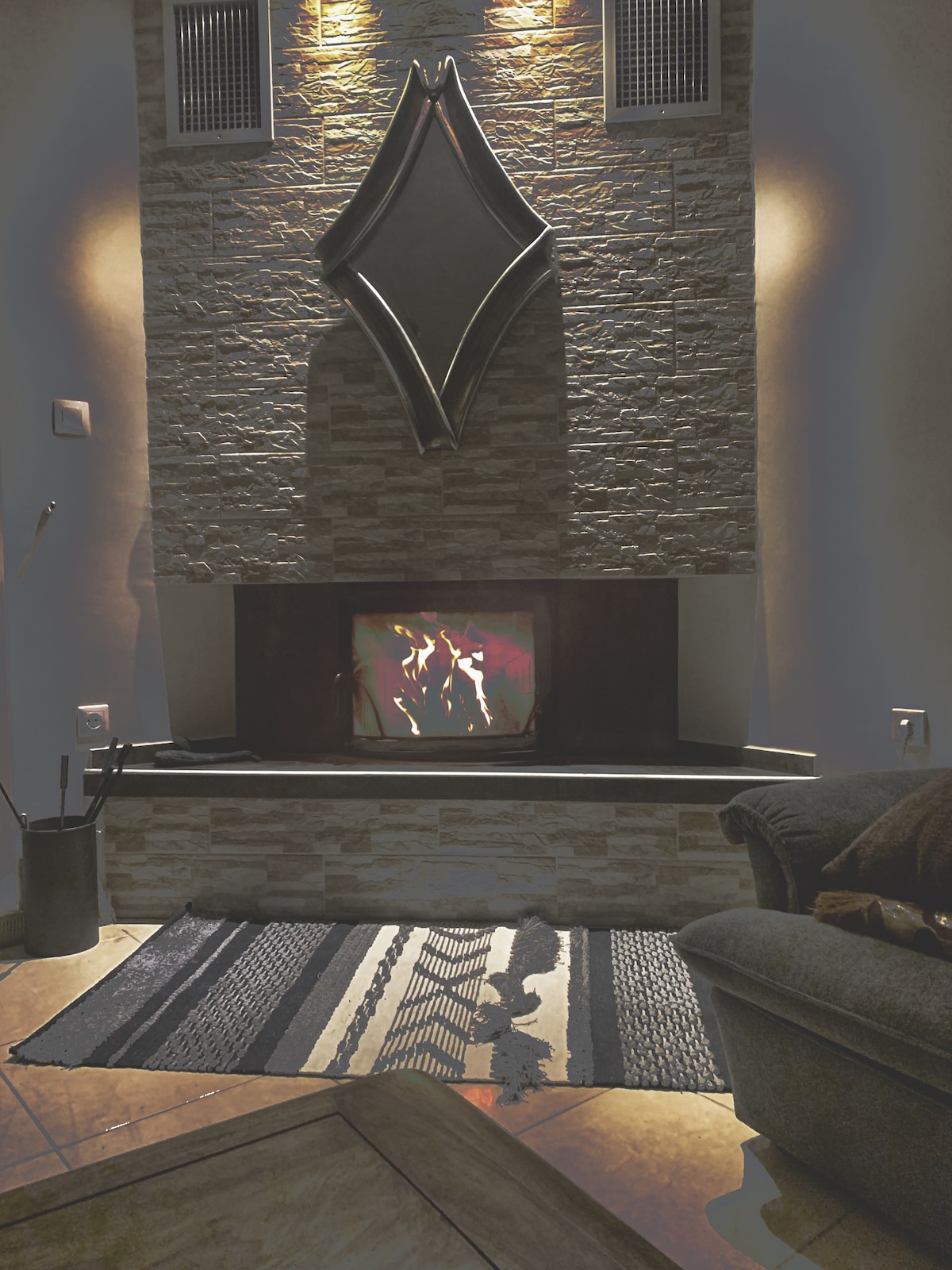 A stone fireplace is centered in the room, featuring a modern mirror above it. A warm fire flickers within, while a textured area rug lies in front. Soft lighting highlights the stone wall, contributing to a cozy atmosphere.
