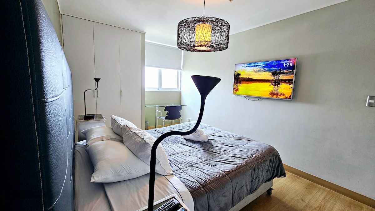 A modern bedroom showcases a spacious bed adorned with soft linens and pillows. A large television is mounted on the wall opposite the bed, while a stylish light fixture hangs above. Natural light enters from a nearby window, complementing the room's neutral color palette.