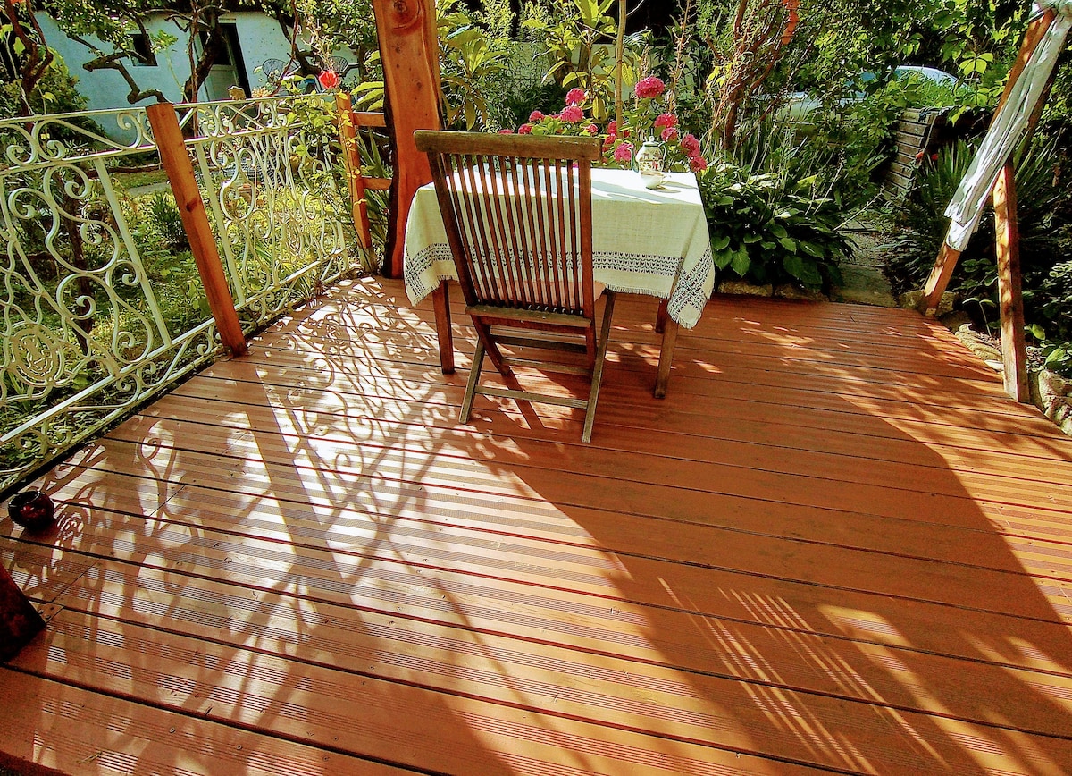 A wooden terrace is presented, featuring a small table set with a light tablecloth and a wooden chair. Vibrant flowers and greenery surround the space, creating a peaceful atmosphere. Soft shadows are cast on the deck from the railing and foliage, enhancing the outdoor setting.