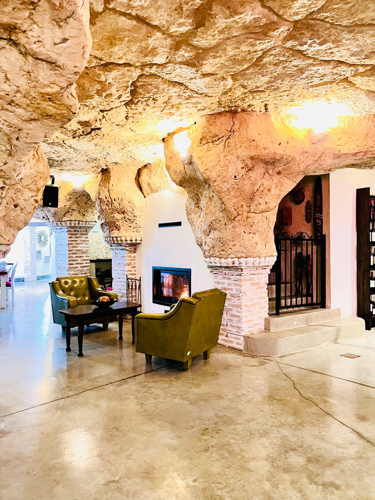 An interior space features natural rock formations, integrating the cave's structure into the design. A cozy seating area includes two green armchairs facing a decorative fireplace. A wooden table is positioned between the chairs, creating an inviting spot for conversation.