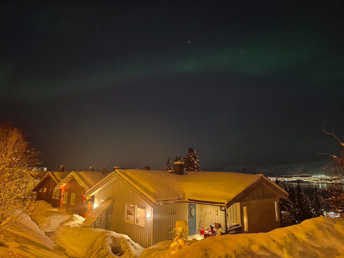 A warm wooden house is highlighted under a night sky, with snow covering the roof and surrounding ground. Glowing lights from the window suggest a welcoming interior, while the northern lights create a soft glow overhead, enhancing the serene winter landscape.