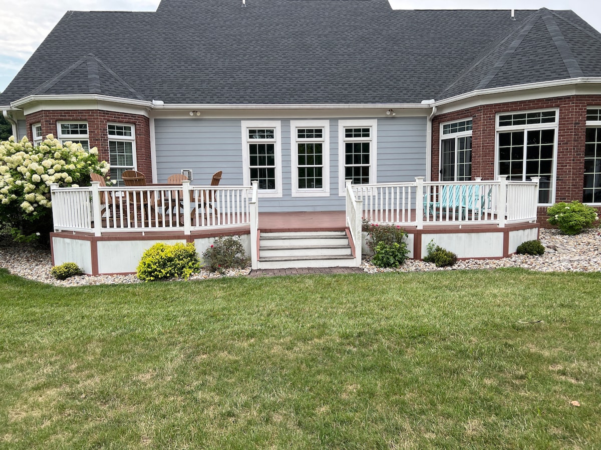 The exterior of a spacious home is shown, featuring a large deck with seating arrangements. Several large windows reflect natural light, framed by manicured landscaping and greenery. A well-maintained lawn surrounds the property, enhancing the serene outdoor setting.